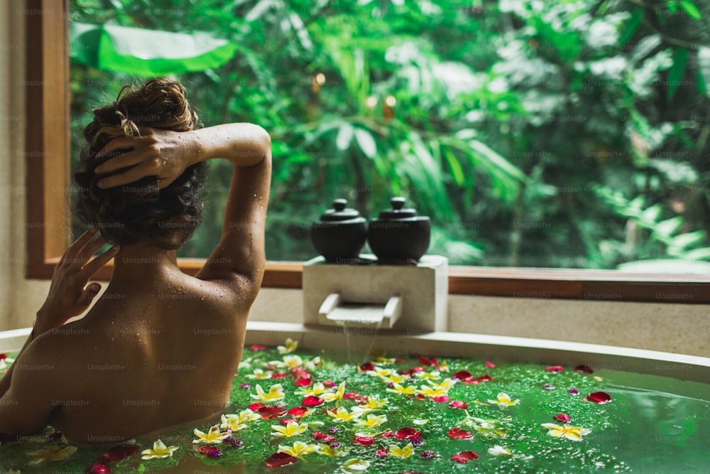 Hermosa mujer joven disfrutando en spa, vista desde atrás. Bañera de piedra de lujo con vista a la selva en ventana. Flores tropicales en el agua. Concepto de tratamiento de belleza.