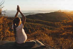 Concept spirituel et émotionnel de l’harmonie avec la nature pendant la maternité. Femme enceinte pratiquant le yoga en plein air sur la colline au coucher du soleil. Vue imprenable sur la montagne d’automne.