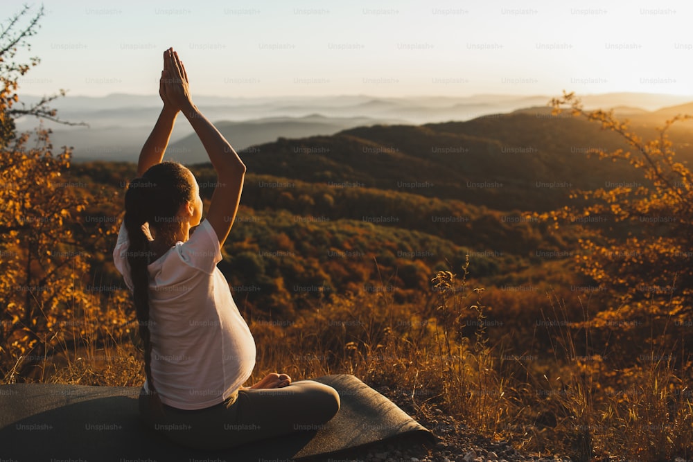Spiritual and emotional concept of harmony with nature in maternity time. Pregnant woman practicing yoga outdoors on hill at sunset. Amazing autumn mountain view.