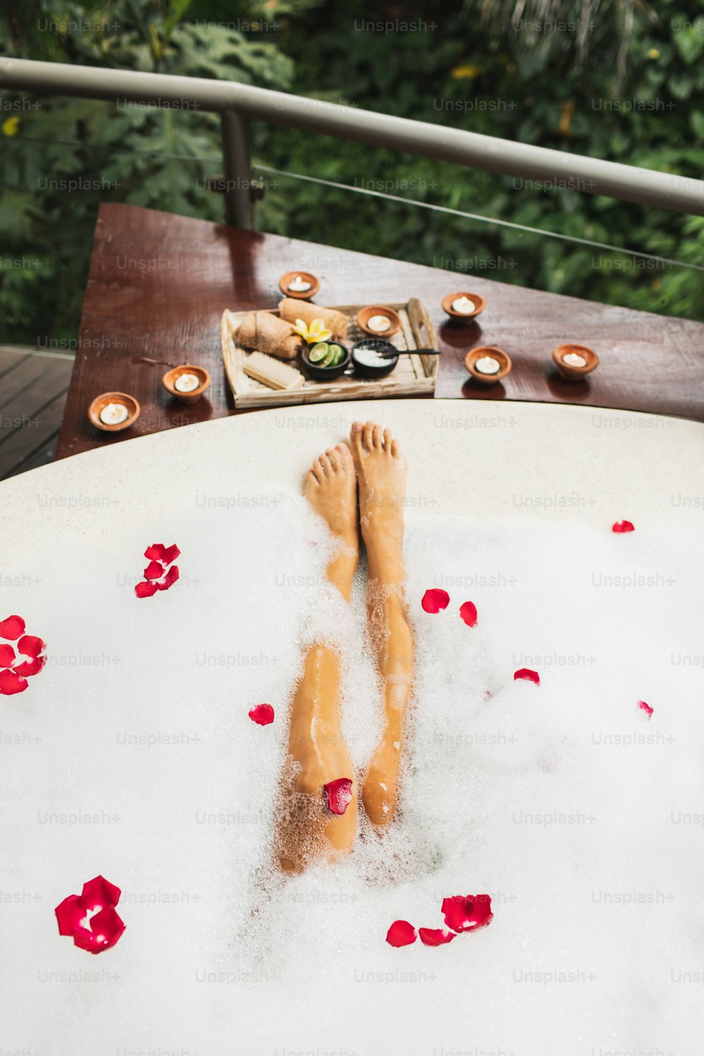 Woman legs in bath tub with foam bubbles and candles around. Romantic spa set with lime, scrub, salt, towels and brush in wooden tray. Organic and natural beauty treatment.