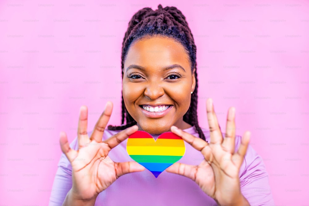 Porträt einer attraktiven hispanischen Frau, die ein LGBT-Regenbogenherz in einem rosa Studio hält.