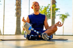man with prosthesis leg practicing yoga sitting in lotus pose on yoga mat meditating outdoors in sun lights.