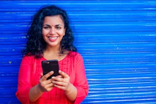 Attraktive schöne glückliche junge lateinamerikanische hispanische Frau mit Teakholz-Bindi auf der Stirn, die auf blauem Wall-Street-Hintergrund lächelt und ein modernes Smartphone hält.