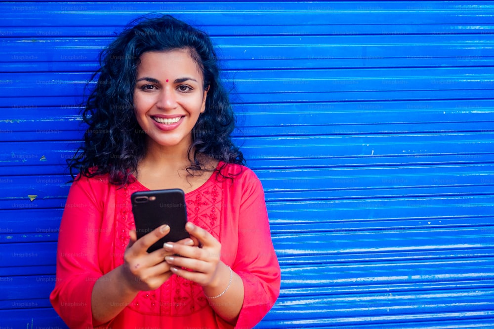 Attractive beautiful happy young latin hispanic woman with teak bindi on forehead smiling on blue wall street background holding smartphone modern.