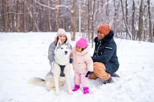 afro man with his caucasian wife having fun with a beautiful daughter playing husky in snowy park.
