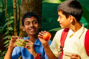 Un écolier indien mangeant un sandwich et son ami avec un sac à dos est assis sur la table et mange une pomme dans le parc.