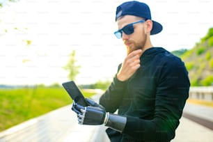 young bearded man with metal arm prosthetic outdoor looking at screen of smartphone.