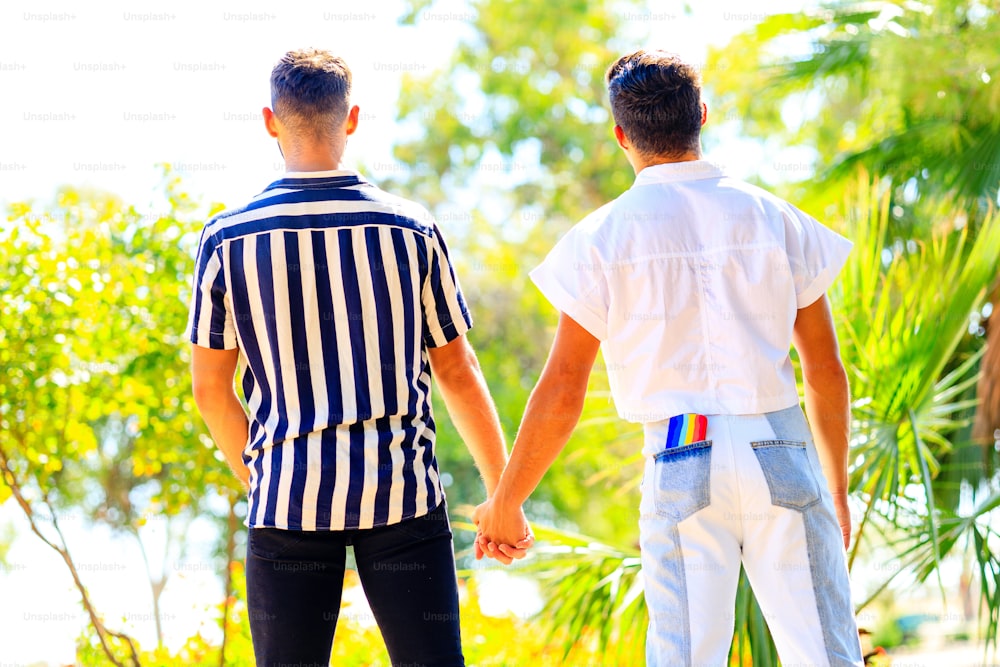young same sex couple in love outdoors holding hands each other and standing back, phone on rainbow case in pocket of jeans.
