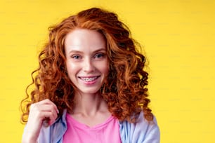 curly redhaired ginger woman with snow white smile brackets on teeth touching a new hairstyle in studio yellow background.