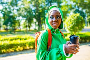 modern afro woman in green hijab with bright make up and piercing nose drinking coffee outdoor and communicating with her friend.