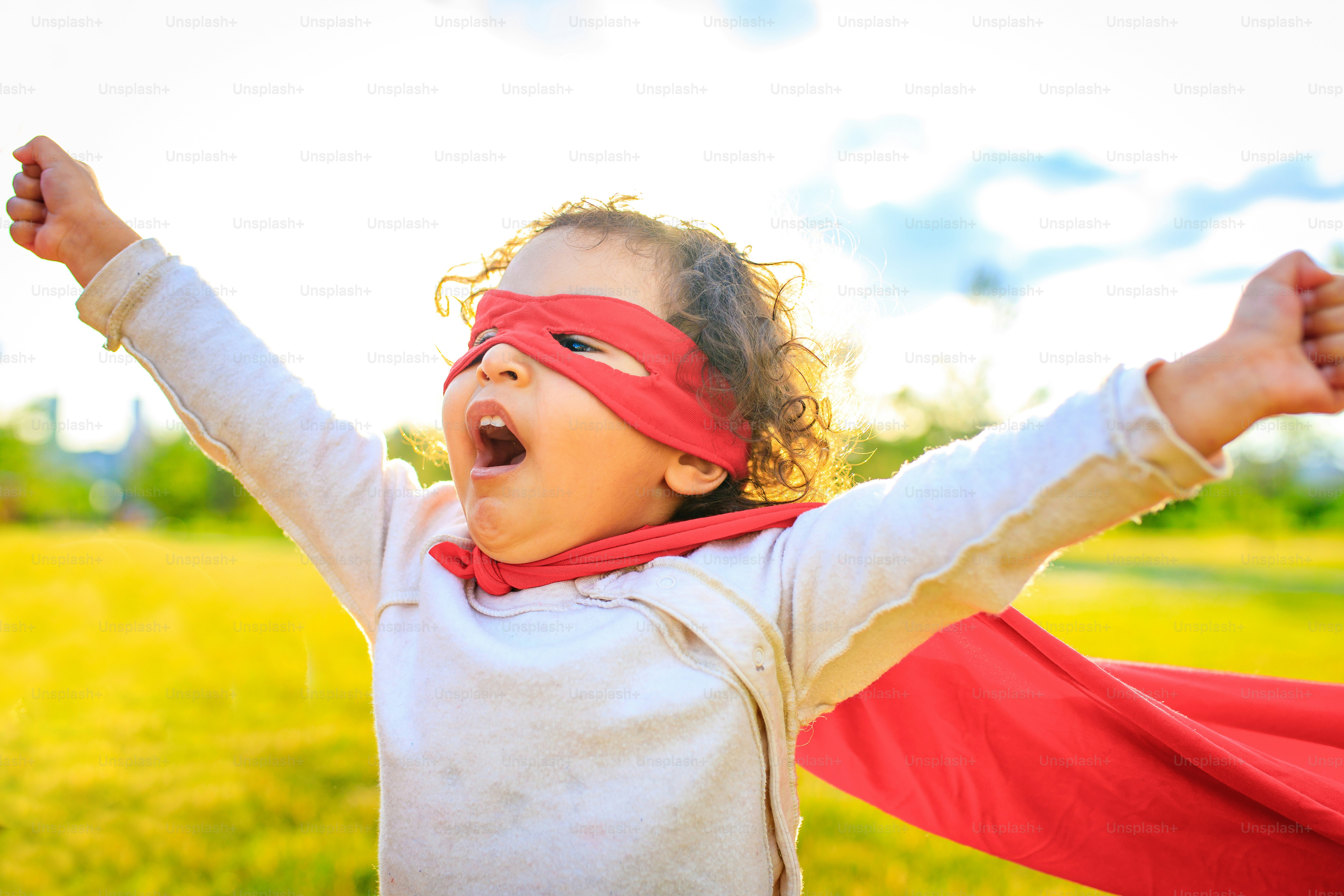 Afroamerikanisches kleines Mädchen in rotem Kostüm und Augenmaske, das einen Helden im Sommerpark spielt.