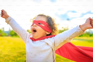 african american little girl in red costume and eyes mask playing a hero in summer park.