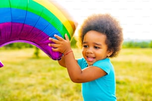 positive mixed race little girl with afro curly hair holding rainbow balloon in summer park.