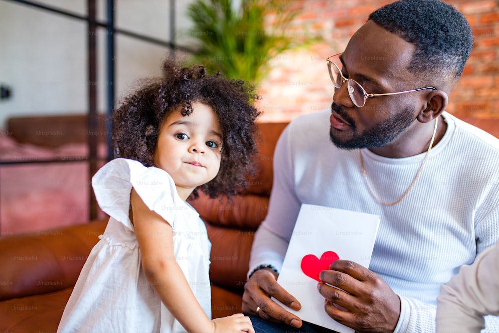 Bebé afroamericano dándole a papá una foto del Día de San Valentín en la sala de estar.