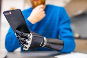 young man with bionic hand hold smartphone while sitting indoors home kitchen background.