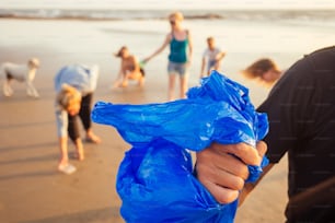 Bel uomo indiano in primo piano che raccoglie la plastica dalla sabbia nella borsa, i suoi amici di razza mista che raccolgono rifiuti sullo sfondo nella spiaggia di Goa India.