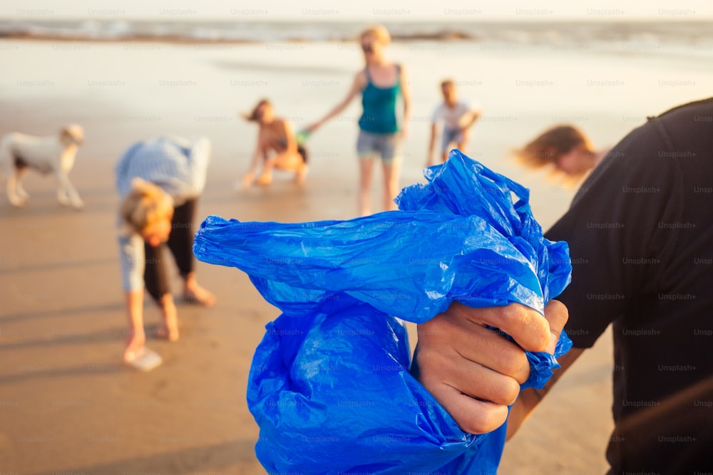 handsome indian man in the foreground collecting plastic from sand into the bag , his mixed race friends picking waste on background in Goa India beach.