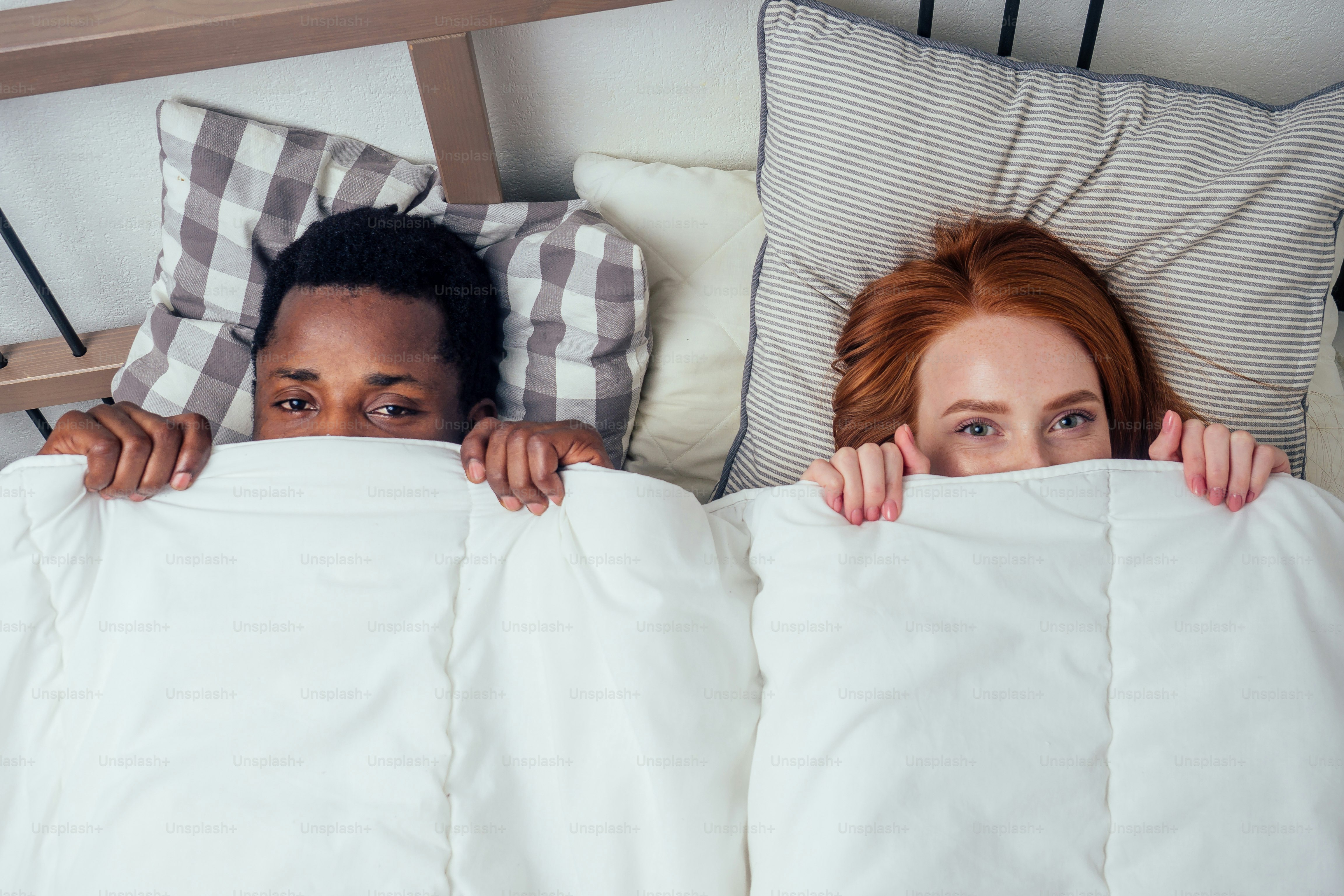 jahe berambut merah, wanita Kaukasia bahagia dan pria Afro multi-etnis bersama-sama, berbaring di tempat tidur, kamar tidur, bersembunyi di bawah konsep selimut.malu.