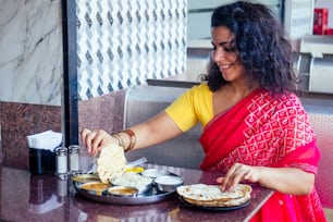 schöne brünette Frau in rotem Sari, die mit Appetit isst, traditioneller Thali Wirh Rise, Quark, Dal im Goa Restaurant Masala Tee.