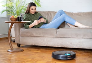 Joven ama de casa hermosa usando robot de limpieza en casa, relajándose en el sofá