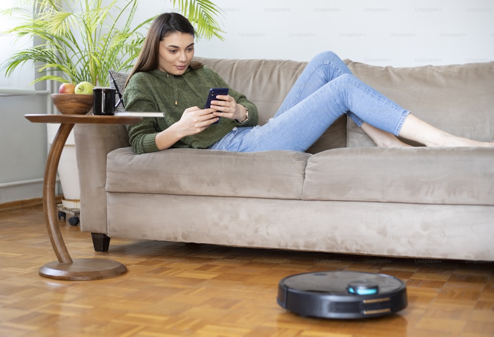 jeune belle femme au foyer utilisant un robot de nettoyage à la maison, se relaxant sur un canapé
