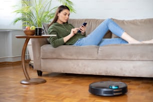 Joven ama de casa hermosa usando robot de limpieza en casa, relajándose en el sofá