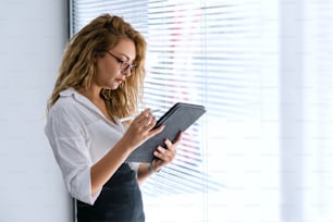 La joven y hermosa mujer de negocios sostiene una tableta móvil en la mano, para revisar su trabajo, mientras está parada junto a las ventanas. Foto de alta calidad