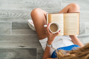 Intellectual young girl sit on floor and drink a cup of coffee, while she is reading a book. High quality photo