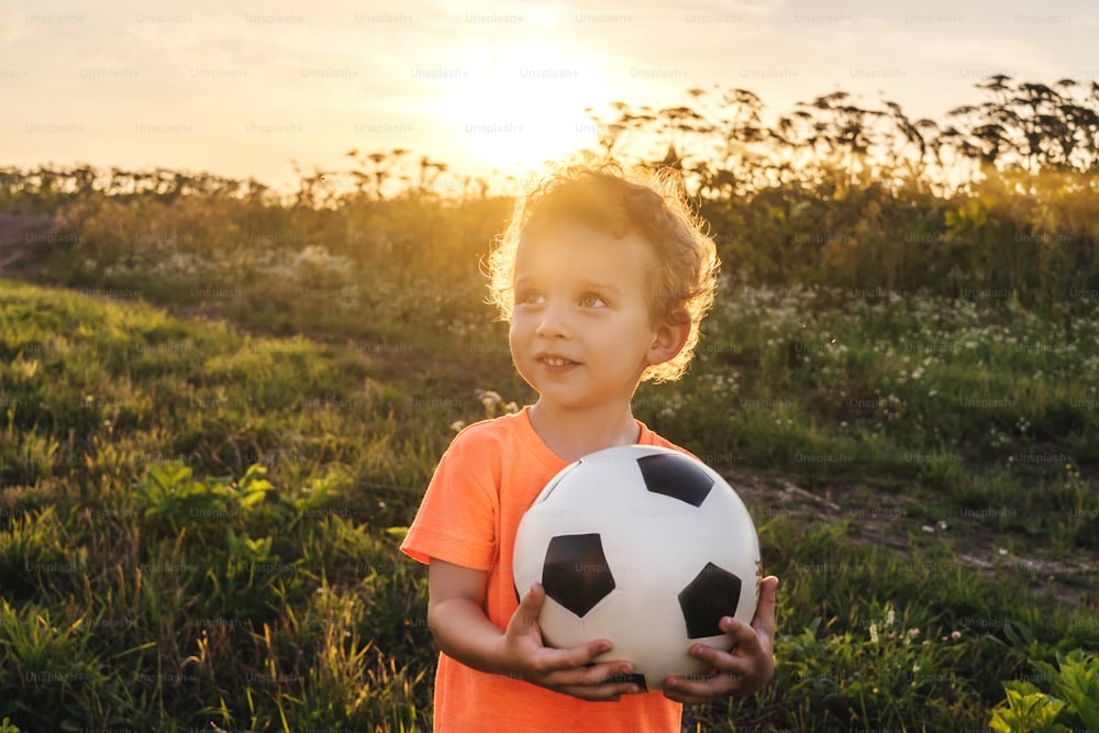 フィールドでボールを持つかわいい巻き毛の少年。背景に美しい夕焼けの光。