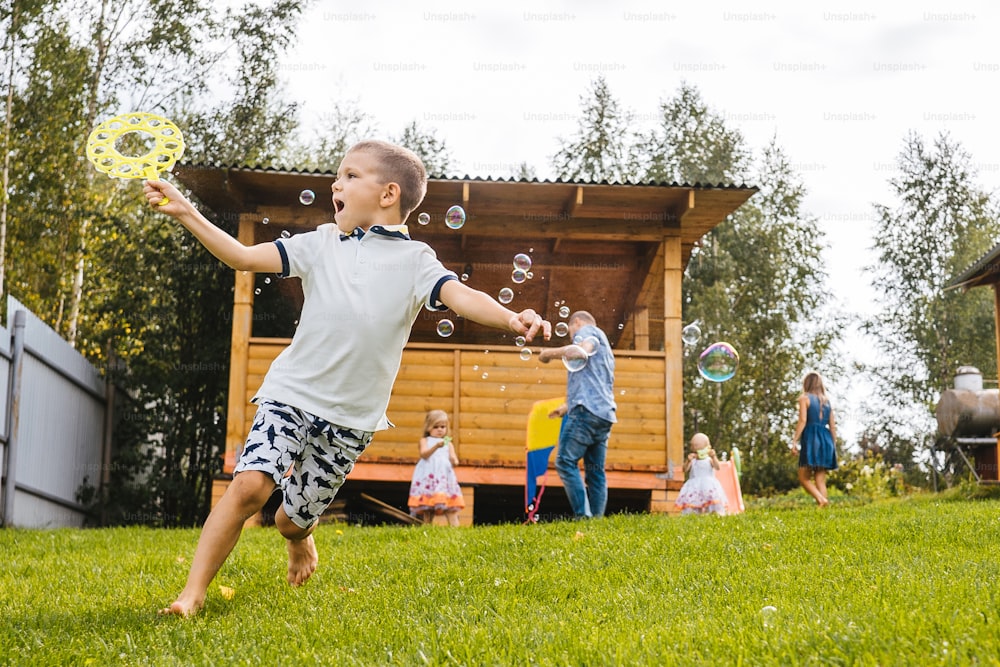 Kleiner Junge rennt mit blasenden Seifenblasen. Hinterhof, Familie im Hintergrund.