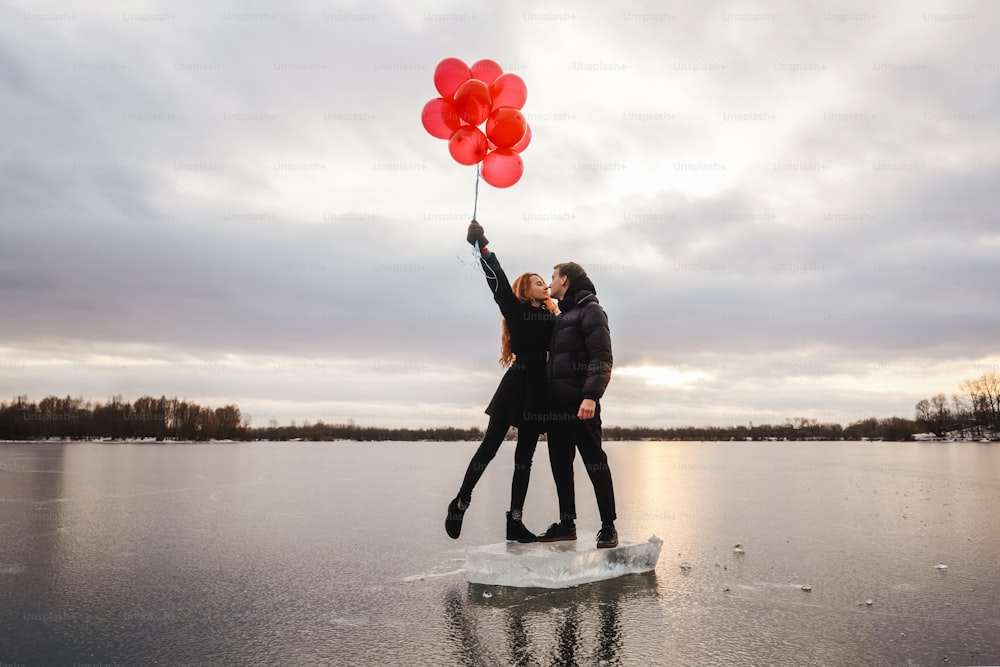 Coppia d'amore con palloncini rossi baciano all'aperto sul cielo e sullo sfondo del fiume di ghiaccio. Luce del tramonto, vestiti casual neri.