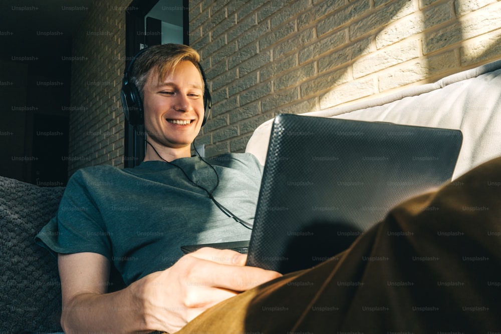 Young smiling man in headphones work with laptop on sofa at home. Brick wall on background, sunny day.