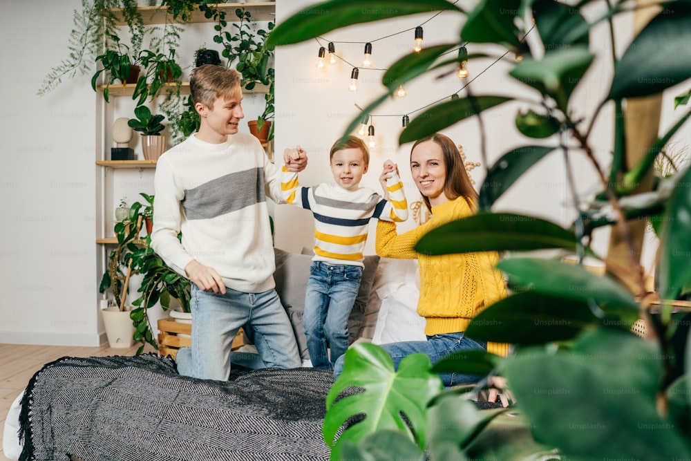 Happy family mother, father and boy have a fun at home in bed. Green houseplants around, sustainable design. Yellow and gray colors 2021 year.