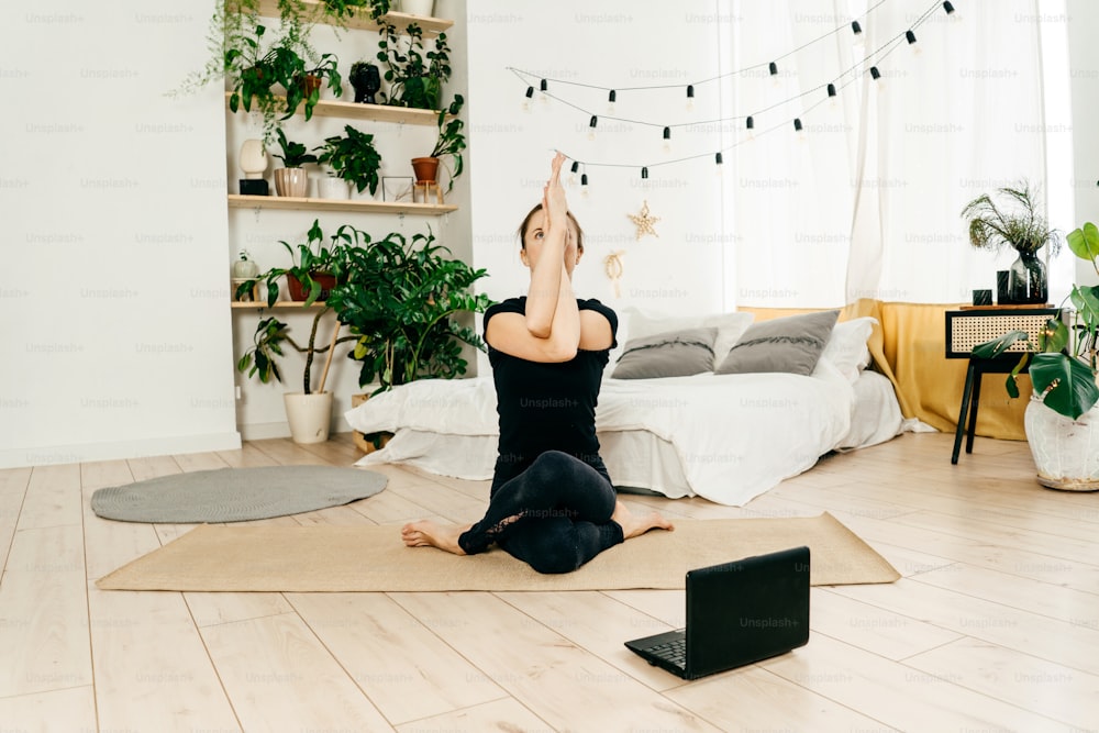 Portrait of beautiful young woman in black sportswear practice yoga at light home. Fitness woman watch tutorial in laptop. Bed and green home plants on background.