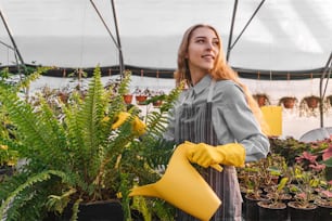 Woman in greenhouse with yellow watering can near fern. Yellow and gray colors 2021.