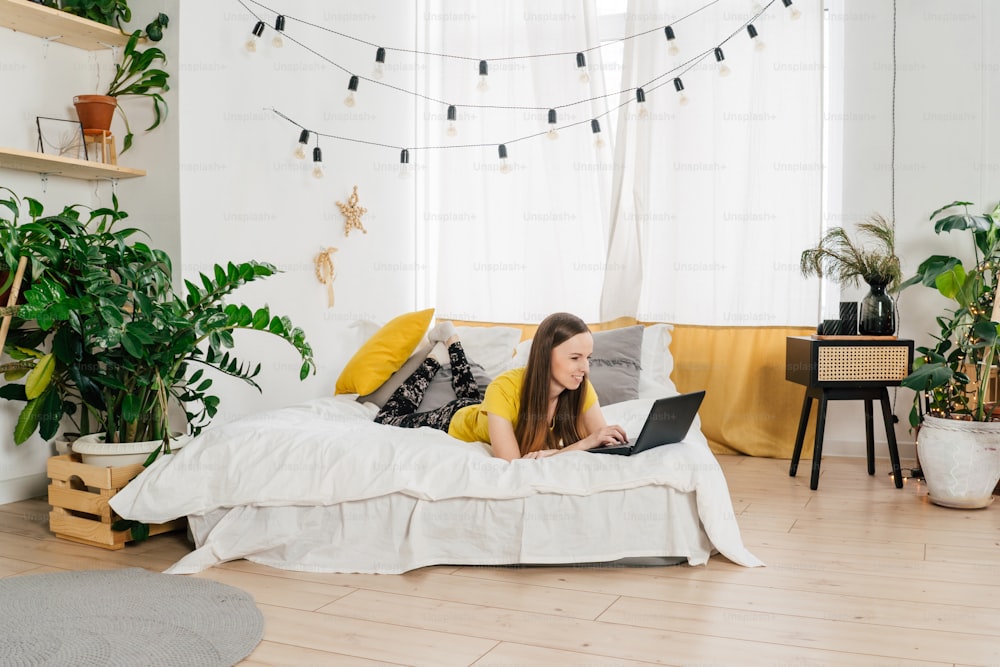 Smiling woman using laptop at home in bedroom. Working from home in quarantine lockdown. Social distancing and Self Isolation. Sustainable design with houseplants.
