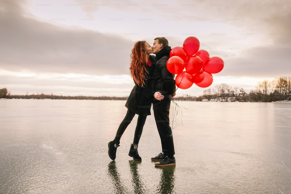 Liebespaar mit roten Luftballons küssen sich im Freien auf Himmel und Eisflusshintergrund. Sonnenuntergangslicht, schwarze Freizeitkleidung.