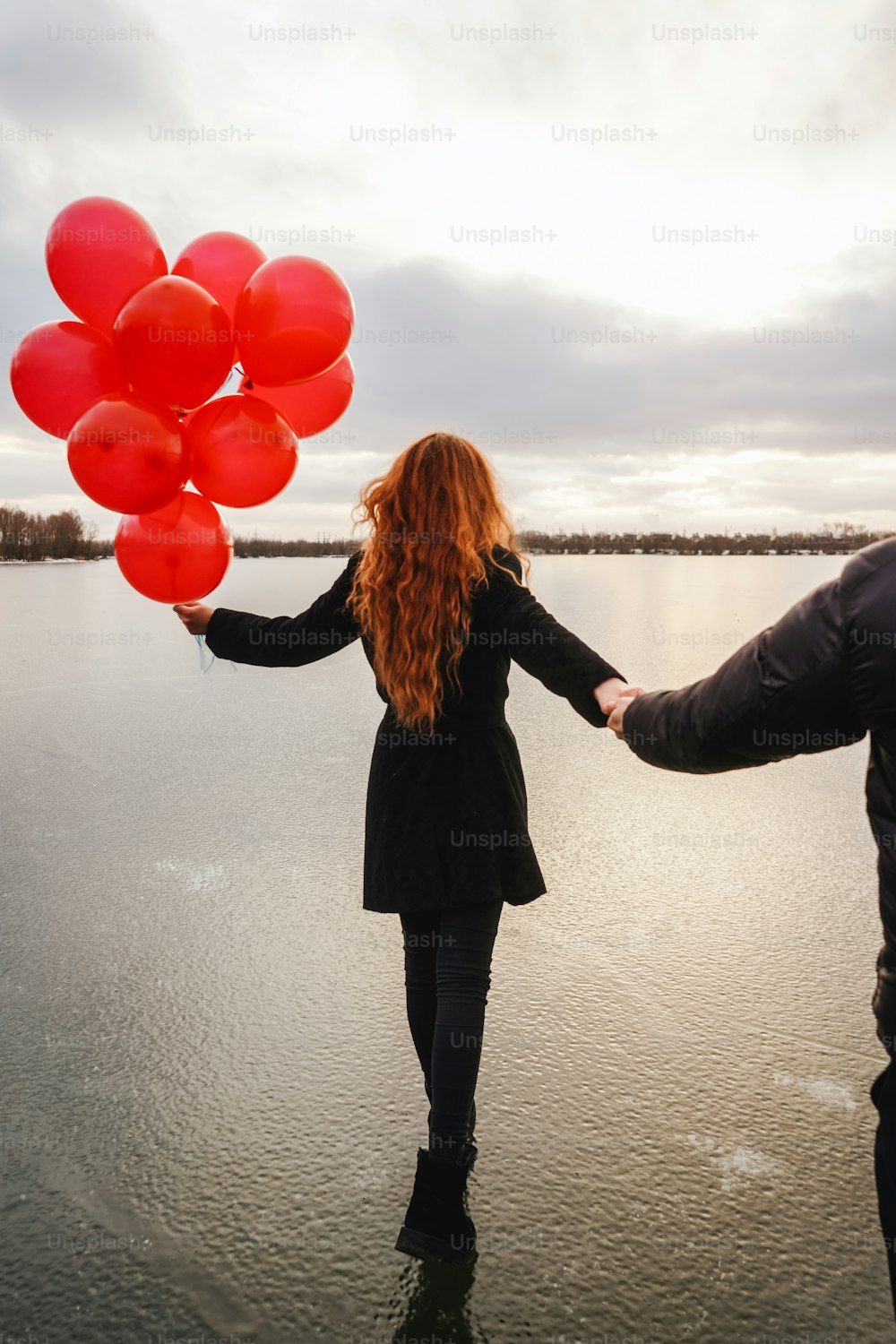 Casal de amor com balões vermelhos ao ar livre no rio de gelo de inverno. Luz do pôr-do-sol, vista traseira. Roupas casuais pretas. Me siga conceito, a garota anda na frente do cara e segura a mão do cara.