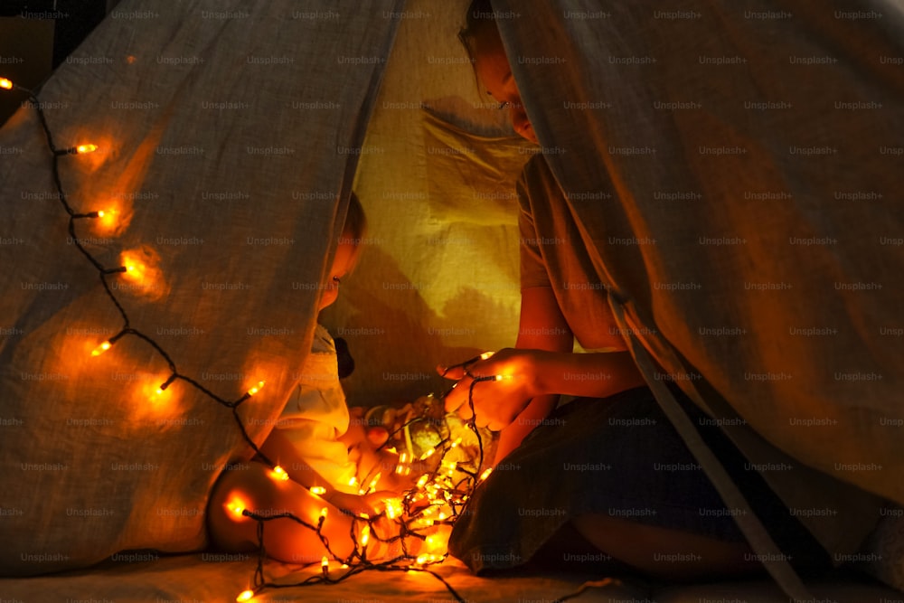 Magic wigwam with garlands and lights with cute girl and her mother inside. Cozy home, dark.