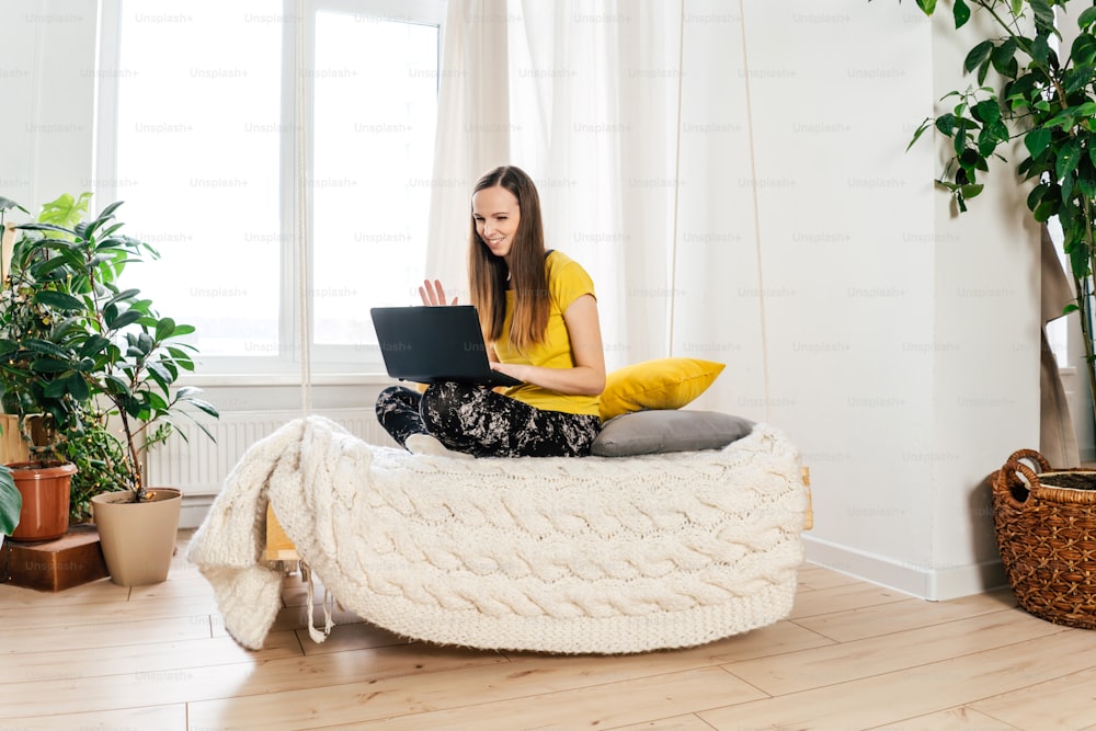Mujer sonriente usando computadora portátil en casa en el dormitorio. Trabajar desde casa en cuarentena encerrado. Distanciamiento social y autoaislamiento. Diseño sostenible con plantas de interior.