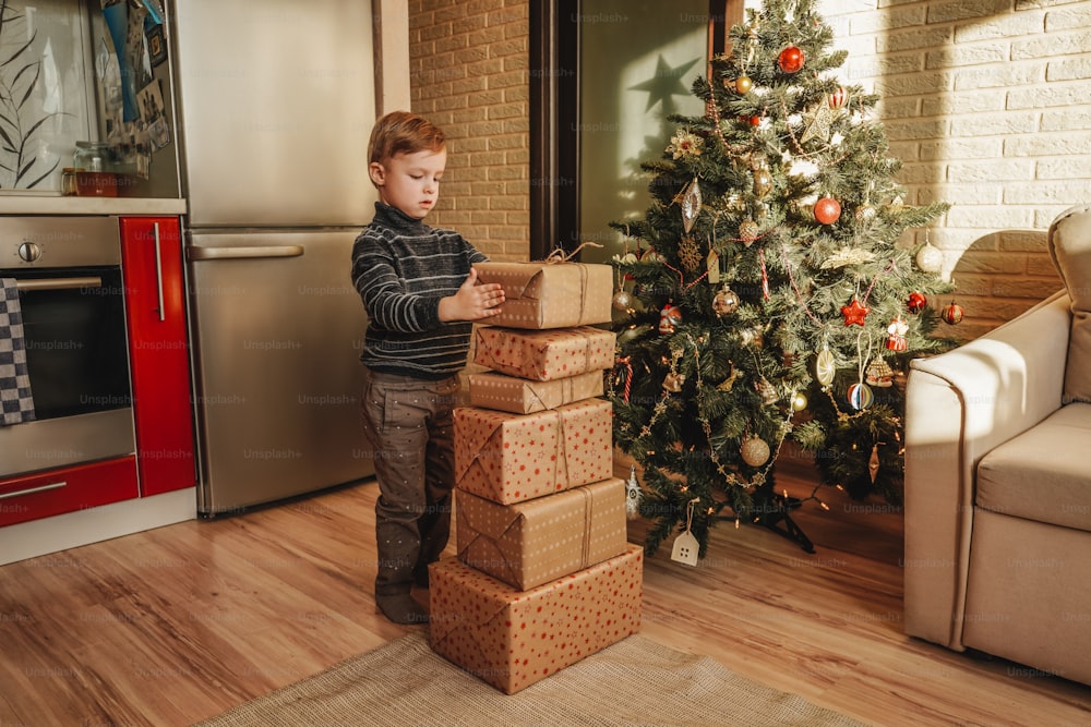 A little boy stand near big stack of Christmas presents. Christmas tree on the background.