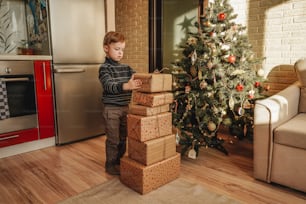A little boy stand near big stack of Christmas presents. Christmas tree on the background.