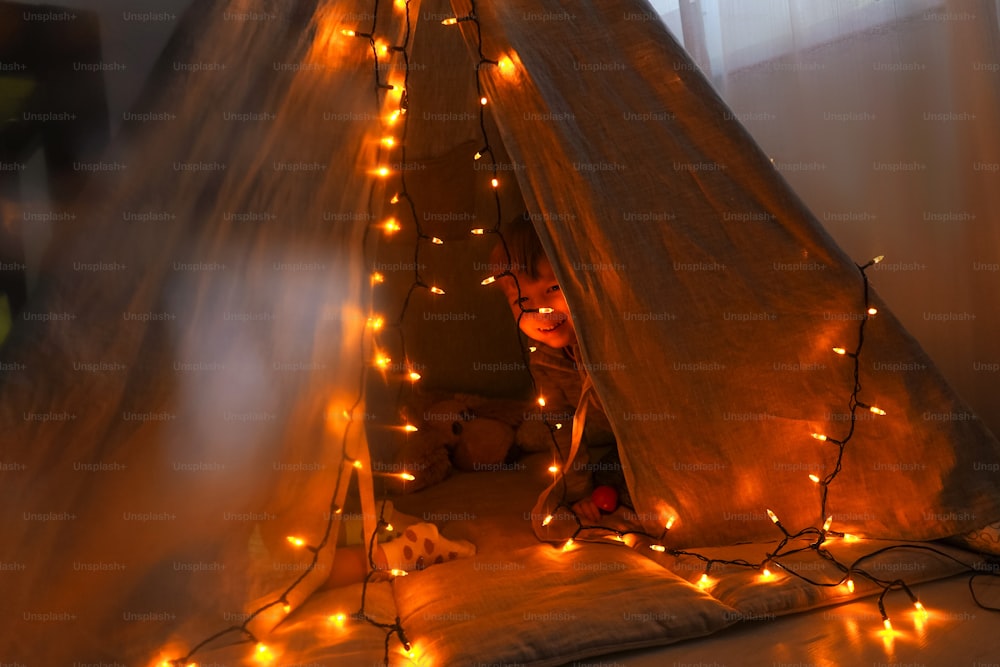 Magic wigwam with garlands and lights with smiling boy inside. Cozy home, dark.