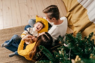 Mamá y papá le hacen cosquillas al hijo cerca del árbol de Navidad. Juego de año nuevo. Vista superior.