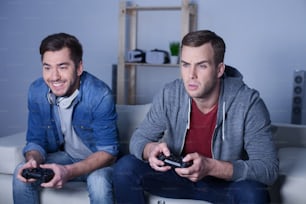 Attractive man is winning his friend in play-station. He is sitting on sofa and smiling. Another man is looking at monitor with seriousness