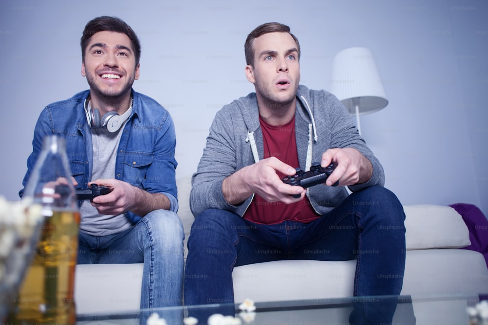 What a great game. Cheerful two friends are playing video games with joy. They are sitting on sofa. The man is smiling