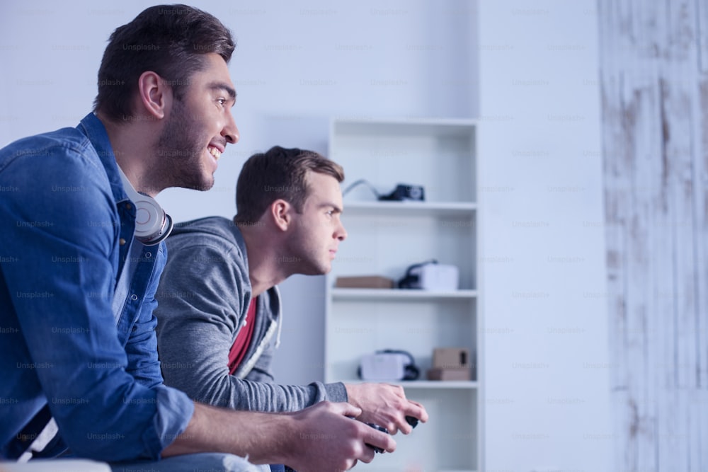 Handsome young men are playing video game at home. They are sitting in profile and smiling. The friends are looking forward with concentration
