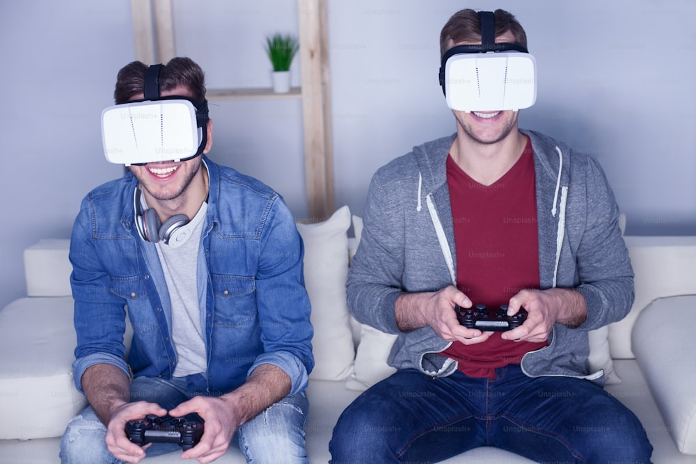 Young men are playing video games at home. They are wearing virtual reality device and smiling. The guys are sitting on sofa