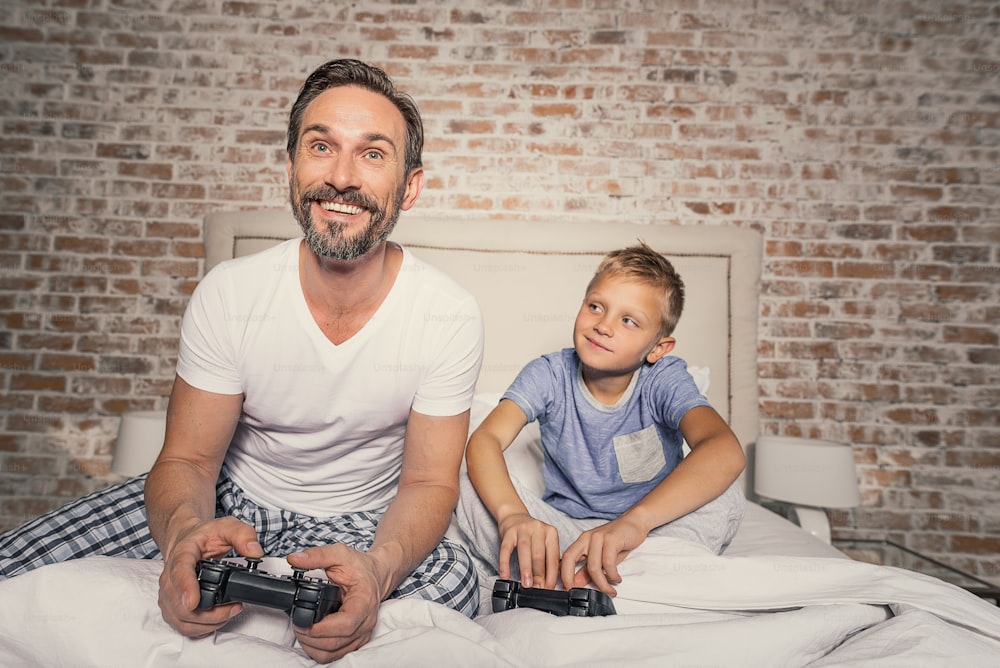 Nunca me ganarás papi. Padre sonriente maduro y su lindo hijo jugando videojuegos juntos mientras están sentados en la cama en la habitación