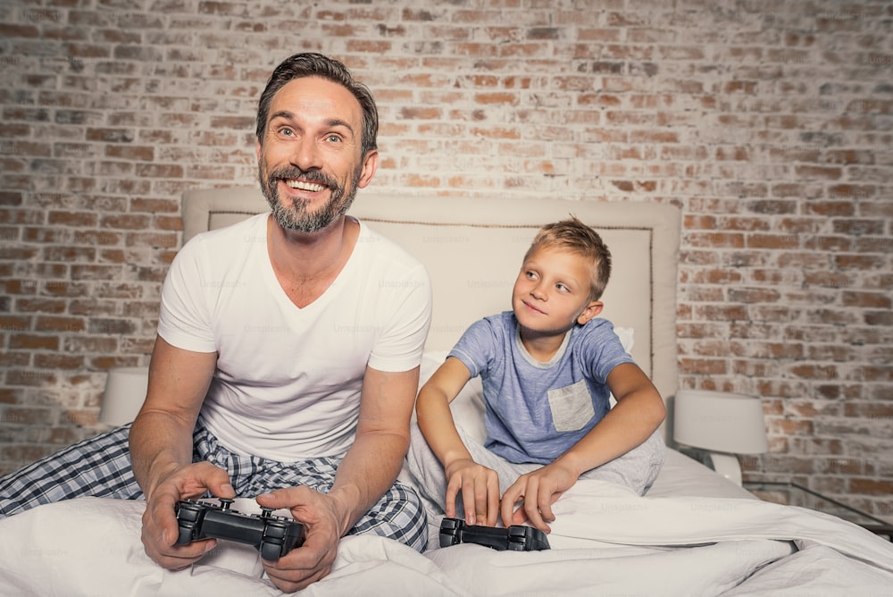 You will never win me daddy. Mature smiling father and his cute son playing video game together while sitting on bed in room